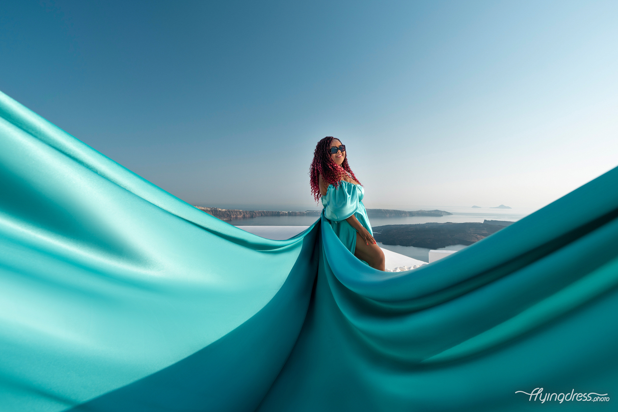 A woman in an emerald green dress poses gracefully with the fabric flowing outward, set against a stunning backdrop of a serene coastal landscape and distant islands under a clear blue sky.